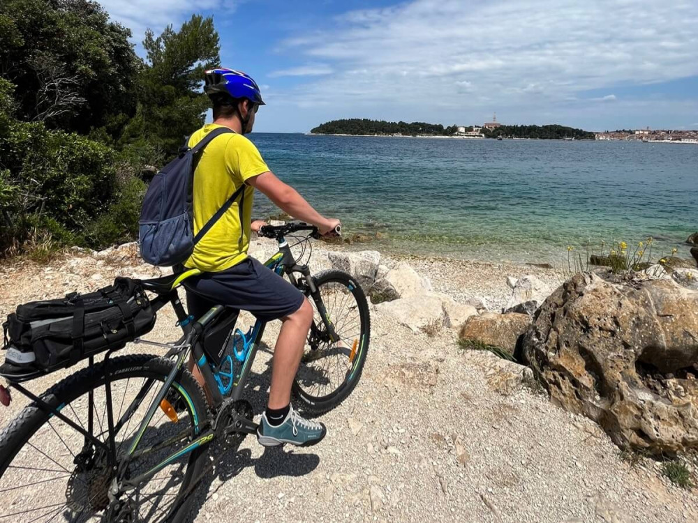 Muž na bicykli na brehu mora na štrkovej pláži s výhľadom na historické mesto.